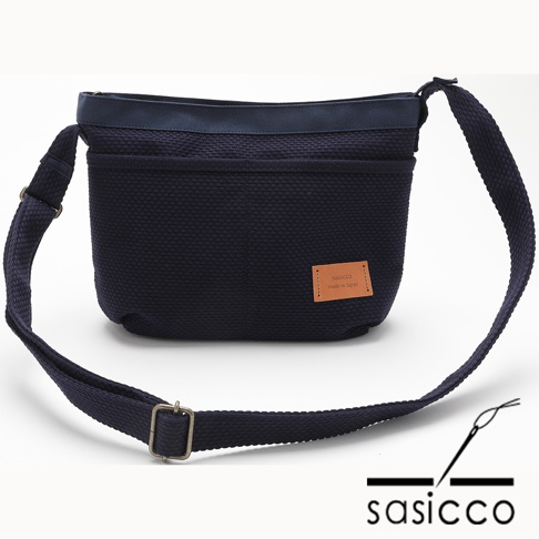 深藍色三河木綿刺子織肩背包，配有可調整的斜挎織帶，包身正面有一個皮革標籤，標示著品牌名稱及產地。