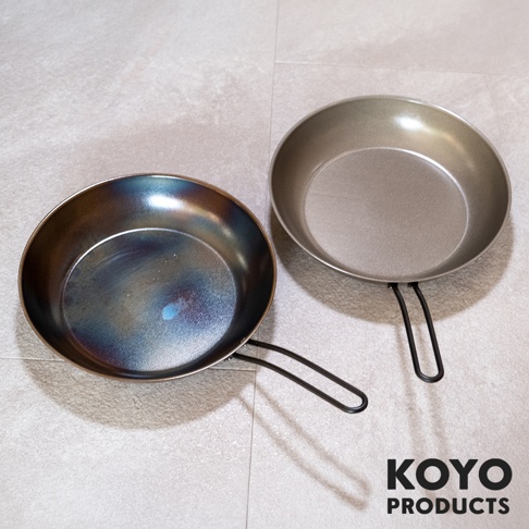 放置於淺色地板上的兩支 KOYO PRODUCTS 淺型鐵製平底鍋，左側鍋身經高溫處理呈現深藍紫色光澤，右側為原色鐵灰質地，兩者均具備壓紋表面與黑色鋼線把手。