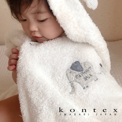 一位穿著白色今治純棉連帽浴巾的嬰兒，浴巾上有大象刺繡圖案，浴巾材質柔軟蓬鬆。