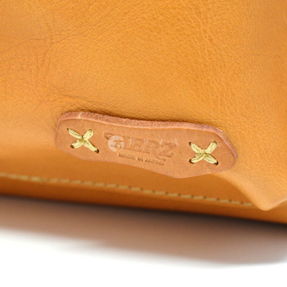 淺棕色皮革後背包特寫，皮革表面紋理細緻，右側有一個縫有黃色線的皮革標籤，標籤上印有日文和英文品牌標誌。