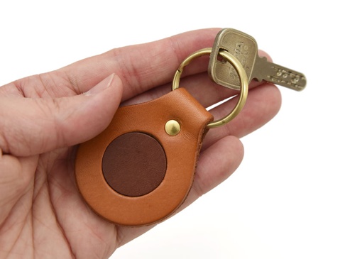 一只手握著一個咖啡色皮革旋轉扣鑰匙圈，鑰匙圈上有一個較深的棕色皮革圓形裝飾，並配有黃銅色金屬扣和一個銀色鑰匙。