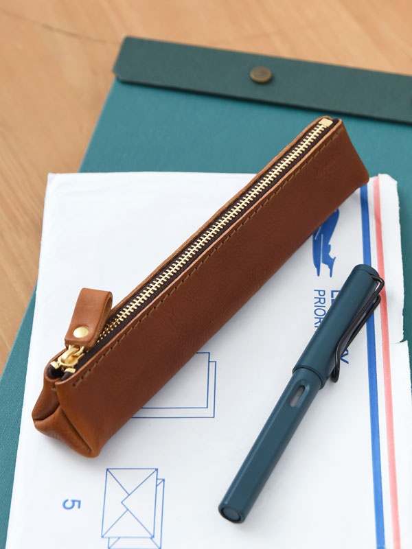 職人手作皮革Organ三角收納筆袋，M尺寸，呈經典棕色，以質感皮革製成，搭配金色拉鍊。旁邊擺放一支深藍色鋼筆，整體置於文件和綠色筆記本上。