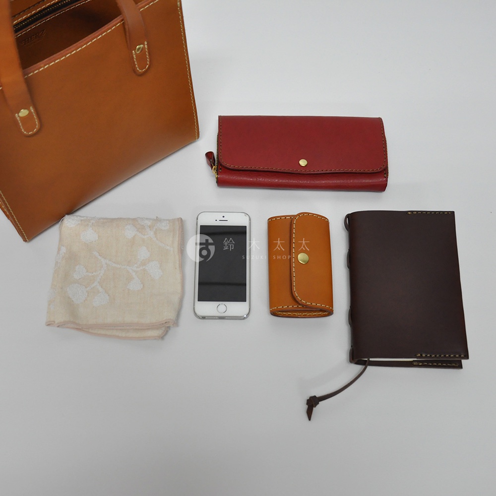 皮革直式托特包（淺棕色）、紅色長夾、白色手機、橘棕色鑰匙包，以及一條淺米色印花手帕，皆為皮革製品，擺放於白色背景上。