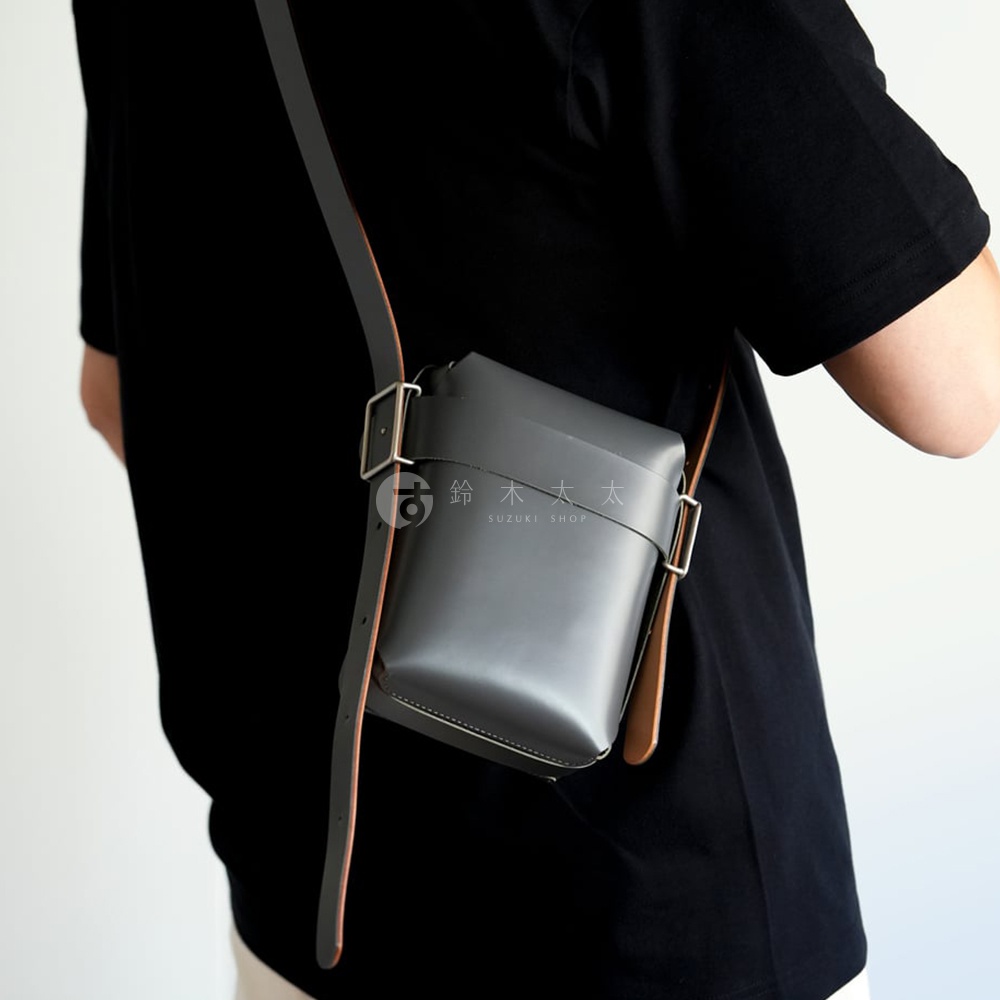 男士身著黑色T恤，斜挎灰色皮革迷你斜背包，包身上印有SUZUKI SHOP字樣。