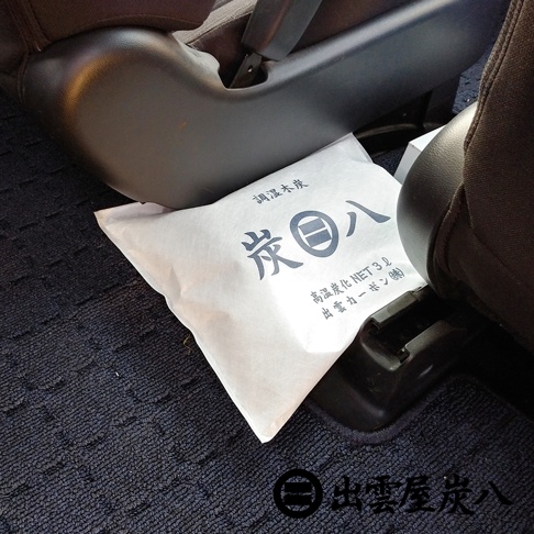 車內方形調濕木炭包，白色包裝印有黑色日文文字，置於汽車座椅底部深藍色地毯上。