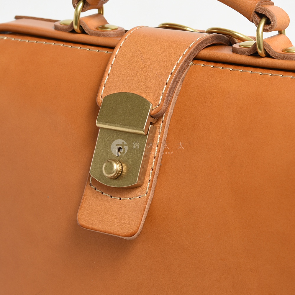 職人手作皮革杜勒斯商務後背包，特寫展示了咖啡色皮革主體、金色金屬鎖扣和皮革提把，縫線清晰，質感細膩。
