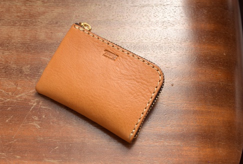 淺棕色皮革製成的L型拉鍊卡片收納夾，表面有細緻紋理，搭配金色拉鍊及米色縫線，置於木紋桌面上。