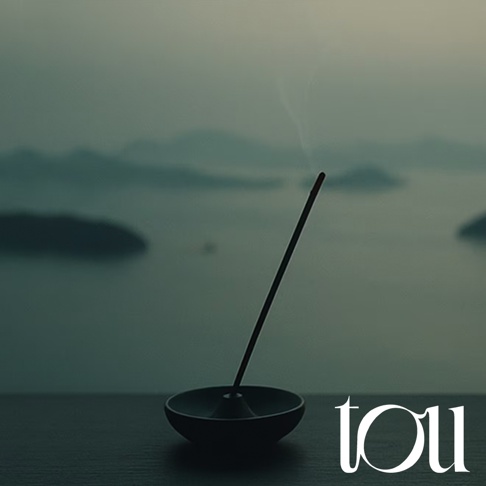 一張靜謐的圖片，深色的線香座放置於水平面上，一支燃燒中的線香筆直豎立，散發出一抹細長煙霧。背景為瀨戶內海的寧靜海景與朦朧島嶼，右下角印有 tou 的品牌字樣，整體營造出沉穩放鬆的氛圍。