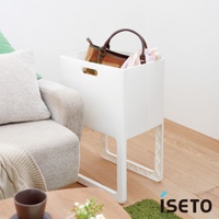 一個白色塑膠折疊高腳置物籃，籃內裝有一個棕色和米色格紋的皮革手提包，以及幾件衣物。置物籃下方有四個白色塑膠支腳，整體擺放在木質地板上，旁邊有灰色沙發和綠色抱枕。