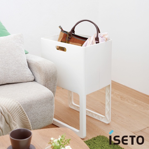 一個白色塑膠折疊高腳置物籃，籃內裝有一個棕色和米色格紋的皮革手提包，以及幾件衣物。置物籃下方有四個白色塑膠支腳，整體擺放在木質地板上，旁邊有灰色沙發和綠色抱枕。