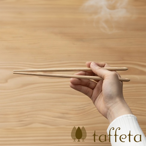 一雙23公分長的楓木圓筷，呈現自然淺木色，由右手持握，筷子末端有細微的品牌壓印，背景為淺色木紋。