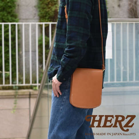 一位身穿綠藍格紋襯衫與牛仔褲的男子，側背著一款日本HERZ手作Latigo硬皮革翻蓋直式單肩包，皮革呈現自然溫潤的駝色，邊緣有明顯的職人縫線細節，設計簡約且具有硬挺質感。
