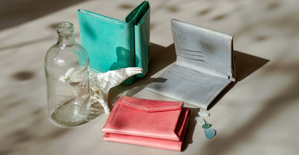 模式B： 一組皮革錢包，包含一個綠色、一個灰色和一個粉紅色，旁邊還有一個玻璃瓶和幾塊海玻璃。