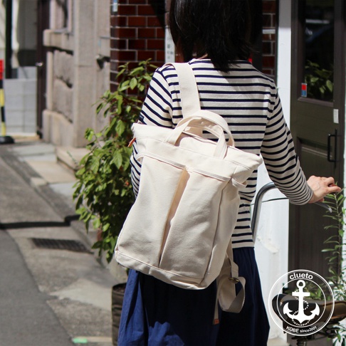米白色帆布後背包，雙肩帶可調節，正面有兩個立體口袋，上方有提把，搭配藍色長裙與條紋上衣，背景為巷弄街景。