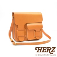 淺棕色皮革製成的厚實單肩包，有雙前袋和翻蓋設計，搭配可調節肩帶。