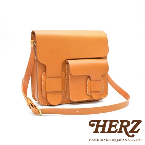 淺棕色皮革製成的厚實單肩包，有雙前袋和翻蓋設計，搭配可調節肩帶。