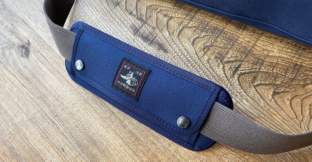 深藍色純綿帆布製成的肩墊，帶有灰色織帶和銀色金屬鈕扣，中央繡有「黑犬印」標誌。