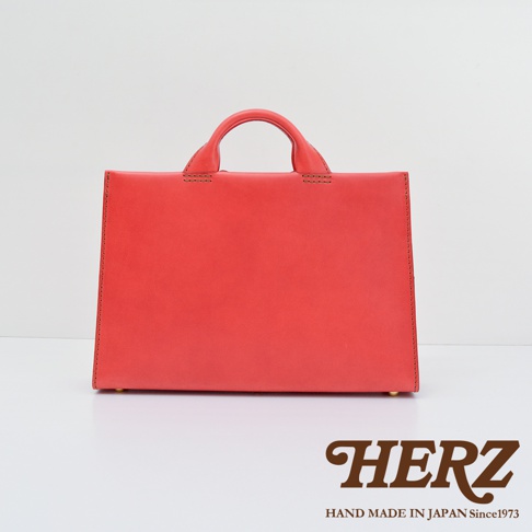 日本HERZ手工皮革紅色公事包，簡約方形設計，頂部設有雙提把，細節處縫線清晰可見，下方印有HERZ品牌標誌及「HAND MADE IN JAPAN Since1973」字樣。