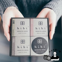 雙手捧著一盒由四個小紙盒組成的香氛火柴禮盒。禮盒外觀為灰褐色，每個小紙盒上印有「hibi」品牌標誌、數字編號（001, 003, 005）及香氛名稱（Lemongrass, Geranium, Tea tree），其中一個圓形紙盒印有「hibi」標誌。