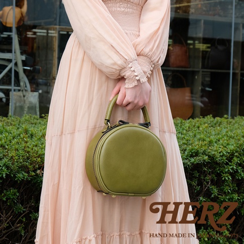 身穿米色長裙的女子手提一個橄欖綠色的圓形皮革包，包包底部有四個金屬腳，包身右下角有HERZ的logo字樣，下方標示著HAND MADE IN JAPAN。