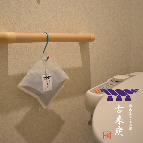 白色半透明包裝袋內裝有黑色木炭顆粒，懸掛於木質掛桿。包裝袋上印有日文標籤。畫面右側為白色馬桶蓋，上面印有紫色繩結圖案及日文文字。