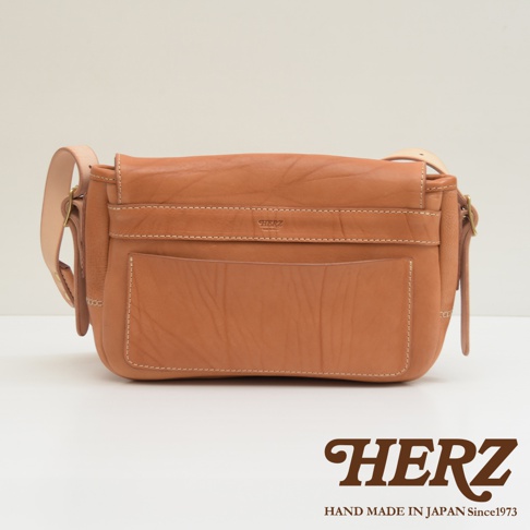 棕色皮革郵差包，正面有方形外袋和翻蓋，帶有品牌標誌和縫線細節，搭配可調節肩帶。