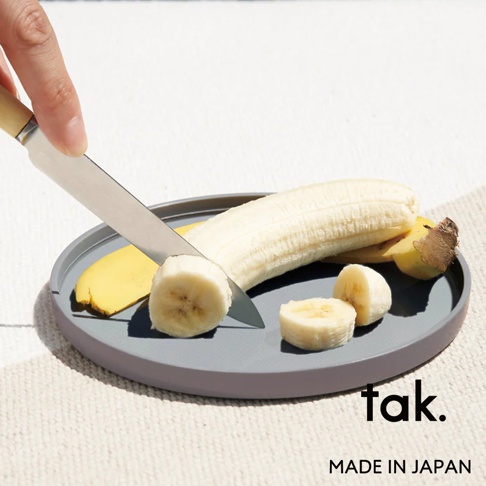 灰色圓形餐盤上，一根剝皮的香蕉正被刀切成香蕉片，香蕉皮置於盤邊。