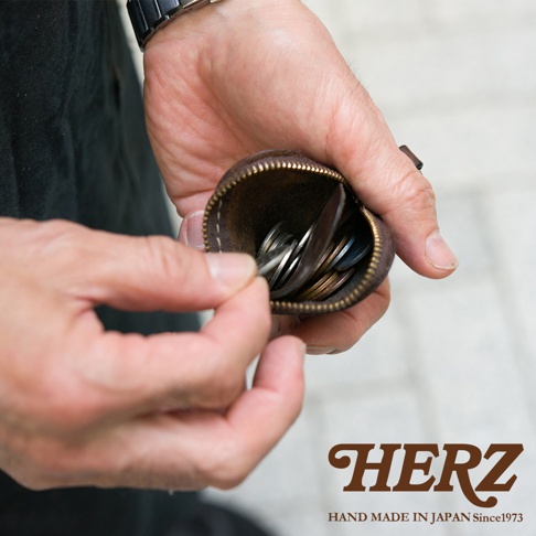 雙手捧著深褐色日本HERZ職人手作皮革零錢包，圓弧形設計以拉鍊開口，皮革質感厚實溫潤。使用者正將手伸入袋中取用內部的金屬硬幣，畫面呈現出細膩的手工藝細節與實用感，右下方標示著HERZ HAND MADE IN JAPAN Since1973字樣。