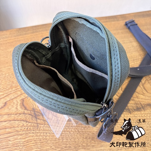 這是一款俯拍視角的橄欖綠色帆布斜背手機包，袋口拉鍊呈敞開狀態，展示內部結構，包含多個分層收納袋與金屬扣環，右下角印有犬印鞄製作所的黑色狼頭與帆布包剪影標誌。
