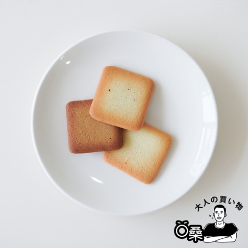 白盤上有三塊方形餅乾，其中一塊較深，另外兩塊顏色較淺，邊緣呈現金黃色。右下角有日文文字和一個卡通人物圖案。