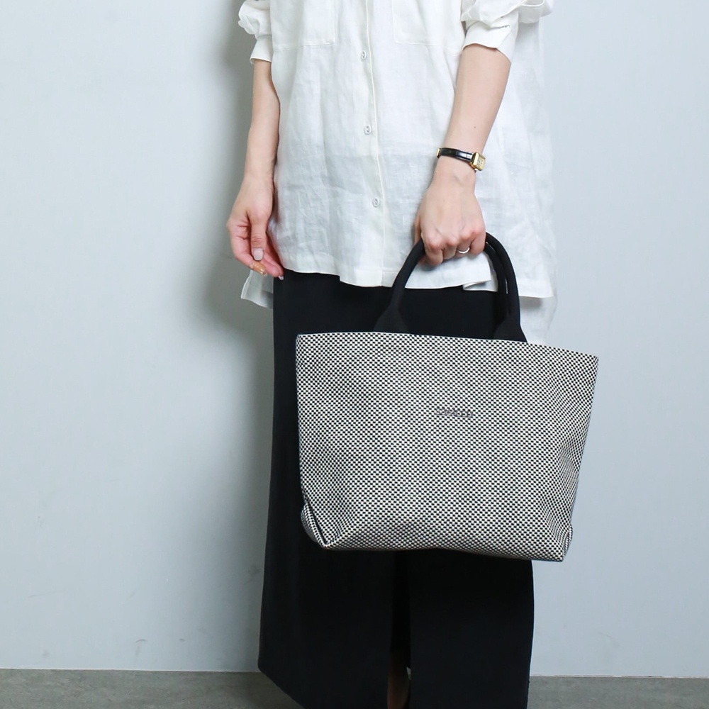 一名女性手持一個黑色和白色格紋的經典托特包，包包由三河木綿刺子織製成，具有簡約的設計和兩個黑色提把。
