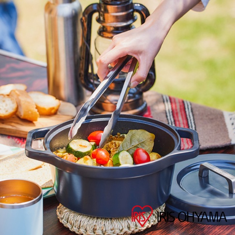 深灰色萬用無水調理鍋，鍋內盛有番茄、櫛瓜、香料等食材，由夾子夾起。鍋旁有鍋蓋、保溫杯及麵包。