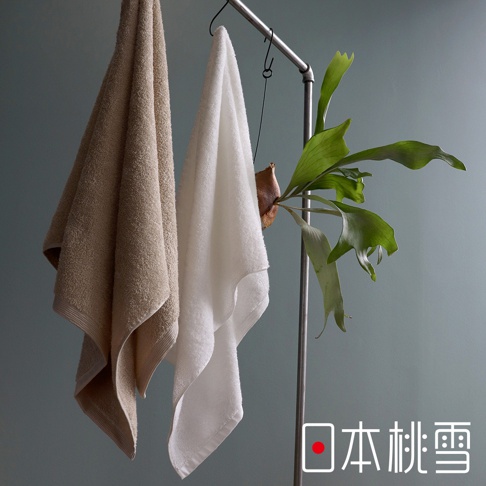 兩條精梳棉浴巾，一條為卡其色，另一條為白色，皆懸掛於金屬架上，旁邊裝飾著鹿角蕨。