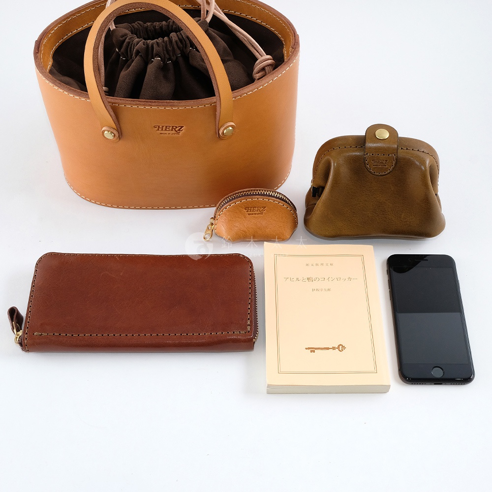 側邊有兩個皮革束口手提包，一個淺棕色，一個深棕色，旁邊還有一個淺棕色和一個深棕色的零錢包。下方有一個長方形的皮革長夾，一本淺黃色的書，以及一支黑色的手機。