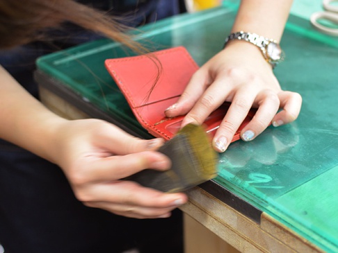 一雙手正在打磨一個紅色皮革三面雙折票券收納夾的邊緣，皮革表面光滑，縫線清晰，收納夾內有隔層。