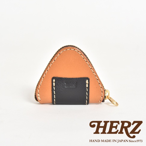 日本職人品牌HERZ手工製作的皮革零錢包，呈現可愛的三角飯糰造型，主體選用淺棕色皮革，搭配黑色皮飾點綴，並以白色粗縫線修飾邊緣，整體採用拉鍊開合設計，右側帶有金屬拉環，品牌Logo壓印於黑色皮革中央。