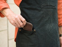 棕色皮革製的拉鍊式零錢包，由手放入黑色圍裙口袋中。