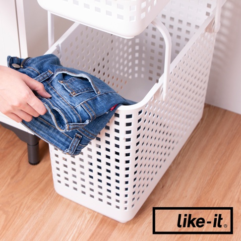 白色塑膠材質的夾縫疊疊洗衣提籃，設計有許多透氣孔，籃身兩側有提把。畫面中，一隻手正將一條藍色牛仔褲放入提籃中。