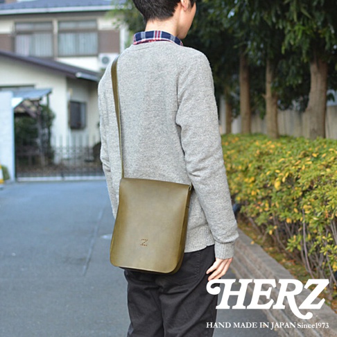 一位男子身穿灰色毛衣和格紋襯衫，肩上斜挎一個綠色皮革包，包上有品牌標誌 HERZ。
