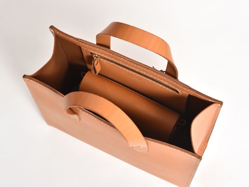 職人手作皮革橫式隨行袋，採用光滑棕褐色皮革製成，設計簡潔，配有兩個提把。袋內有一個拉鍊隔層，以及一個可見的長形皮革配件。