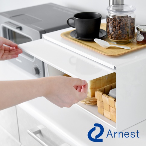 白色金屬廚房小烤箱置物架，上方擺放著黑色咖啡杯、咖啡豆玻璃罐與果醬小罐，架子下方收納著麵包和編織籃。