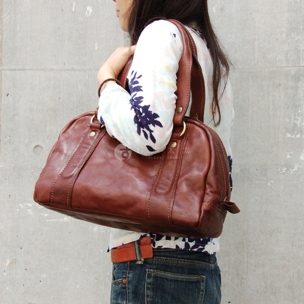 職人手作皮革水洗加工迷你波士頓包，呈現深棕色，材質為柔軟皮革，包身有雙提把及金屬拉鍊，搭配印花上衣與牛仔褲。