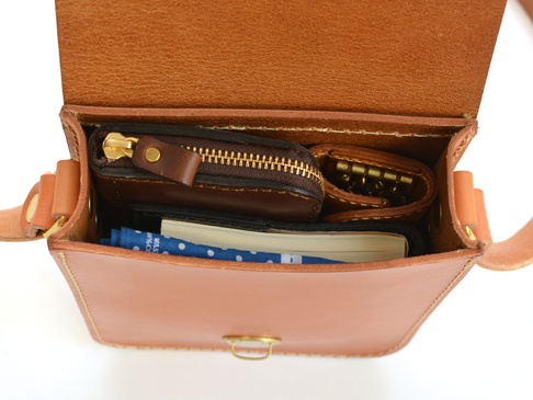 職人手作皮革經典皮扣單肩包 (CP-97)，打開的淺棕色皮革包內，可見一個深棕色拉鍊零錢包、一個金色鑰匙圈，以及一本筆記本和藍色印花布。