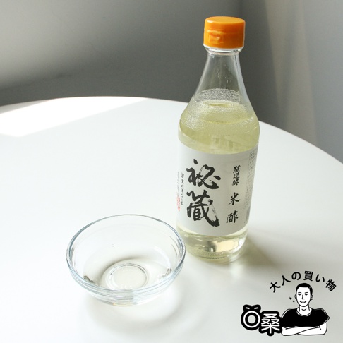 玻璃瓶裝的秘藏米醋，醋液呈淡黃色，瓶蓋為橘黃色。旁邊放有一個透明玻璃小碗，碗中盛有少量米醋。
