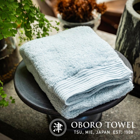 淺藍色純棉毛巾，柔軟蓬鬆，帶有細緻的條紋織邊，置於黑色陶瓷盤上，背景為綠色植物和裝飾。