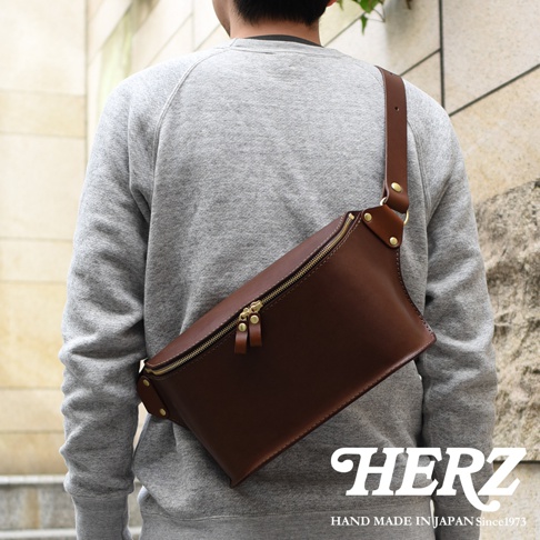 咖啡色皮革立體斜背包，採用職人手作工藝，配有金色拉鍊與金屬扣環，以斜背方式展示於穿著灰色連帽衫的模特兒身上。