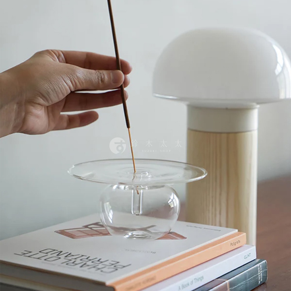 一隻手正將一根線香插入一個透明玻璃製成的線香座，線香座底部是圓球形，上部是一個扁平的圓盤，圓盤上有水滴。後方是一個蘑菇造型的檯燈，旁邊有書籍堆疊。