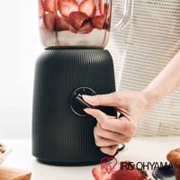 黑色直條紋紋理的果汁機/料理機，搭載透明果汁壺，壺內有切片草莓與奶昔。一隻手正在操作機身上的黑色旋鈕，旋鈕上有「P」、「O」、「I」等標示。