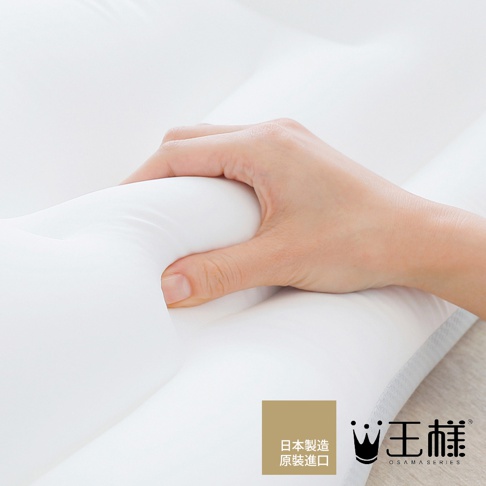 左手輕壓白色極夢枕，枕面呈現柔軟的凹陷，質感光滑。枕頭邊緣有淺灰色網狀織物。右下角有「日本製造 原裝進口」及「王樣 OSAMASERIES」的品牌標誌。
