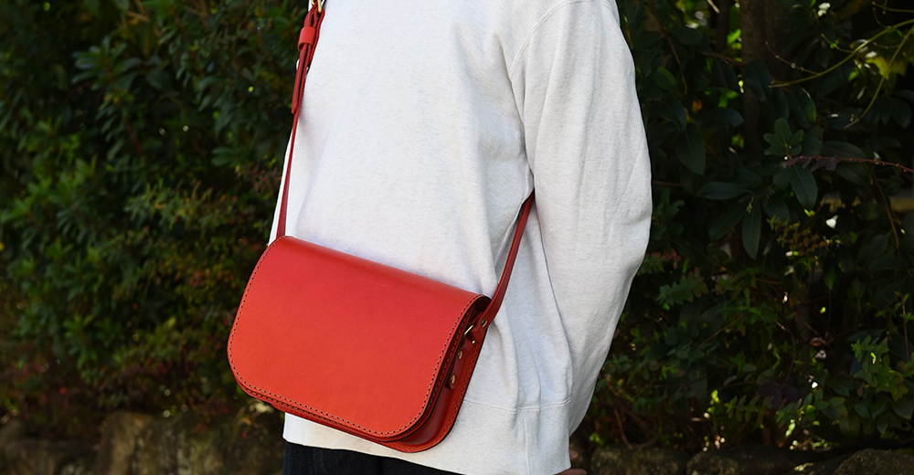 紅色皮革翻蓋橫式單肩包，配有可調節肩帶，包身縫線清晰，風格簡約。